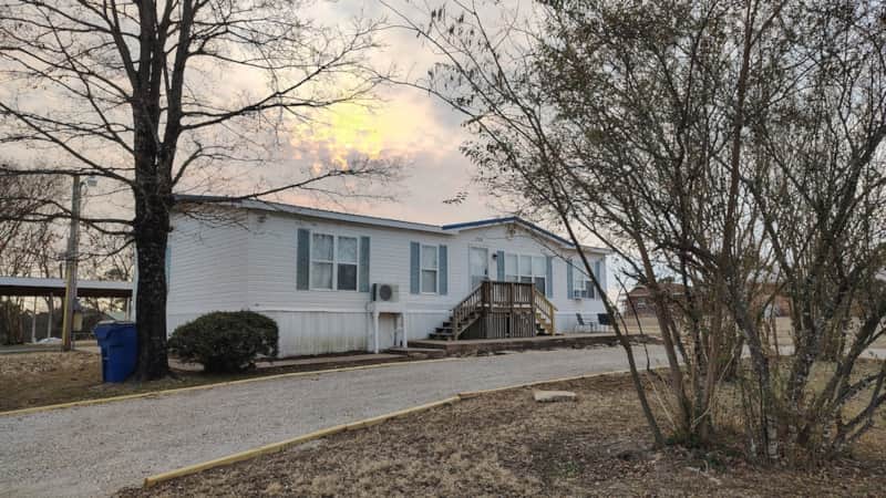 Exterior view of the Lichner Resort Stays manufactured home with white siding, blue shutters, and front wooden deck at dusk in Salesville Arkansas