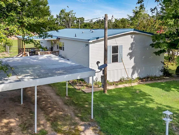 Elevated view of a white manufactured home with a blue metal roof, covered deck, and a freestanding white metal carport in the grassy yard at Lichner Resort Stays.