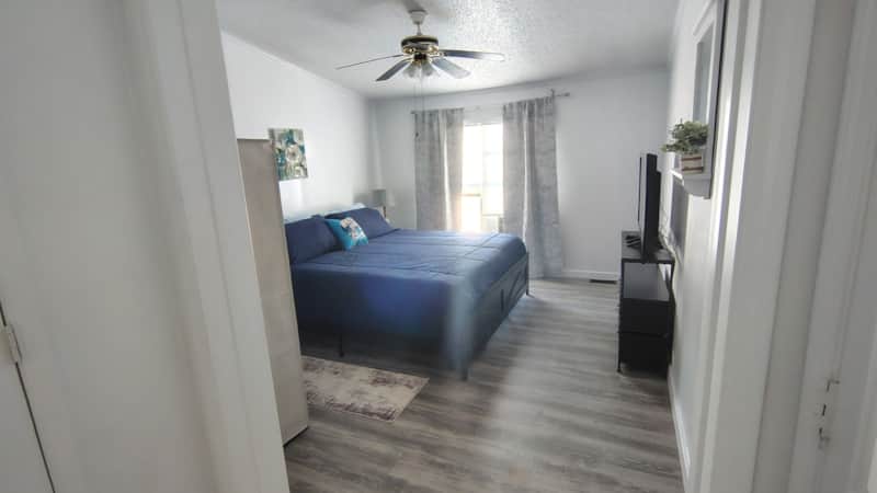 Primary King Suite bedroom viewed through doorway with navy bedding, wardrobe, ceiling fan, and wood-look flooring at Lichner Resort Stays